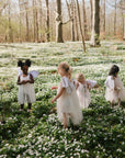 Four children in white fairy costumes walk through a sunlit forest, giggling as they clutch mushie Snack Cups amid blooming white flowers and tall spring trees.