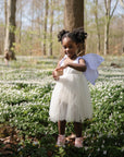 A young girl in a white dress smiles in a sunlit forest, clutching mushie Silicone Baby Food Containers among white wildflowers, with trees and soft light in the background.