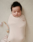 A newborn with dark hair is swaddled in a beige blanket on a matching ribbed surface. Beside the baby lies the mushie Silicone Pacifier Clip | Eva, a food-grade silicone pacifier accessory.