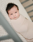A baby with dark hair lies on the mushie Mini Muslin Crib Sheet in light beige, gazing up with wide eyes. The breathable 100% cotton muslin sheet and crib rails are visible in the foreground.