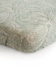 A close-up of the mushie Mini Muslin Crib Sheet, made from 100% cotton muslin with a light green and cream paisley pattern, fitted snugly around a mattress corner.