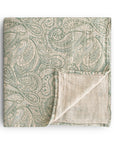 The mushie Organic Cotton Muslin Swaddle Blanket in Green Paisley is folded to display its patterned cream side and plain underside, neatly arranged against a white background.