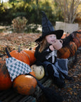 A young toddler in a witch costume smiles outdoors among pumpkins, holding a mushie Snack Cup. Autumn leaves, a black witch hat, cape, and broom complete the festive Halloween scene.