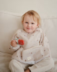 A young child sits on a light-colored couch, smiling and holding a red popsicle while wearing the mushie Long Sleeve Bib with small sailboat patterns.