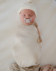 A newborn, swaddled in beige and wearing a matching hat, sleeps peacefully on a white surface using the mushie Silicone Pacifier Clip | Eva to keep their pacifier secure. Made from food-grade silicone, this accessory ensures safety and style.