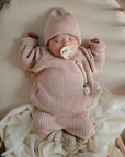 A baby sleeps on a cream blanket in a crib, wearing mushie Chunky Knit Pants made from organic cotton, a matching pink knit hat, and a pacifier clipped to their sweater.