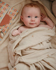 A smiling baby with light skin and reddish hair lies on a soft towel, wrapped in a beige rainbow-patterned blanket next to a colorful pouch, wooden toys, and mushie Cradle Cap Brushes with gentle silicone bristles—perfect for bath time.