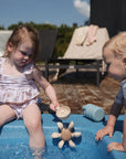 Two young children play by a small blue pool. One girl in a ruffled swimsuit pours water into the mushie Water Wheel Bath Toy as a boy in swimwear watches. Pool toys and a lounge chair can be seen in the background.