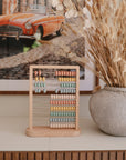 The Mushie Wooden Abacus, featuring pastel-colored beads, rests on a wooden surface beside a large textured vase with dried plants and a framed photo of a vintage orange car in the background.