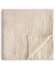 The mushie Organic Cotton Muslin Swaddle Blanket in the Kites color is shown folded, with one corner flipped up to display both sides of the soft baby blanket.