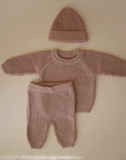 The mushie Chunky Knit Pants are part of a cozy three-piece baby set in light brown organic cotton, featuring a long-sleeve sweater, matching hat, and pants, all displayed flat against a beige background.