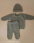 A mushie Chunky Knit Pants set in soft gray organic cotton, including a long-sleeve sweater and beanie hat, is displayed flat on a neutral beige background.