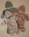 Three mushie Chunky Knit Pants sets—each with pants, sweater, and hat in pastel green, pink, and beige—are neatly arranged on a beige surface. All are cozy knitted baby outfits made from organic cotton.