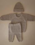 The mushie Chunky Knit Pants are shown in beige organic cotton as part of a baby outfit with a long-sleeve sweater and beanie, all laid flat on a neutral background.