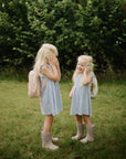 Two young girls with long blonde hair wearing matching blue striped dresses, rubber boots, and backpacks stand on the grass outdoors, smiling as they hold mushie Phone Press Toys to their ears. Green bushes in the background invite sensory exploration.