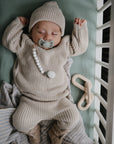 A baby sleeps in a crib with a green sheet, wearing mushie Chunky Knit Pants and a beige knitted outfit. A pacifier with a beaded clip is attached to their sweater, and a soft toy rests nearby.