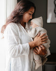 A woman in a white shirt gently holds a sleeping baby in beige knitwear near a bright window, soothing the infant with a FRIGG Butterfly Anatomical Silicone Pacifier 2-Pack (0-6 Months) by FRIGG.