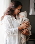 A woman in a white shirt gently holds a sleeping baby dressed in mushie Chunky Knit Pants, with soft daylight shining through the window for a peaceful, tender scene.
