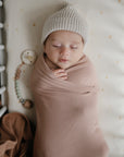 A baby sleeps peacefully in a crib, wrapped in a beige swaddle and wearing a knitted hat. Next to the baby, the mushie Silicone Pacifier Clip | Eva lies on the star-patterned mattress as an adorable pacifier accessory.