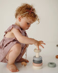 A young child in a mauve dress crouches on a white background, stacking pastel rings from the mushie Everyday Play Gift Set ($48 value), encouraging fine motor skill development.