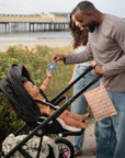 A man offers the mushie Daisy Rattle Teether to a baby in a stroller, while a woman stands nearby on a beachside path with green bushes and a pier in the background.