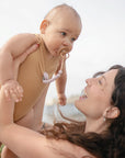 A woman with long brown hair smiles while lifting a baby outdoors. The baby wears a tan bodysuit and uses a FRIGG Daisy Natural Rubber Pacifier from the FRIGG 2-Pack collection. The background is softly blurred.