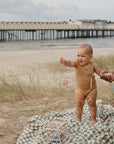 A smiling baby in a brown onesie stands on a checkered blanket at the beach, holding an adult’s hand for support, with the mushie Silicone Pacifier Clip | Hera attached. A pier with yellow buildings appears in the background over the water.