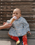 A baby in a blue patterned outfit, red socks, and a mushie Long Sleeve Bib sits on a wooden bench, licking a pink ice cream cone held by an adult.