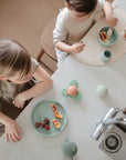 Two young children sit at a kitchen counter with snacks on light plates, enjoying mealtime together as mushie Suction Spinner Toys provide a playful, sensory experience nearby.