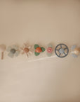 Six pastel-colored, flower-shaped and circular mushie Water Wheel Bath Toys are lined up horizontally on a beige surface, casting faint shadows.