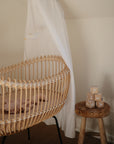 A light wooden bassinet with a white canopy stands beside a small wooden stool holding stacked beige blocks. The mushie Moon Lovey Blanket, made from organic cotton, adds comfort to the softly decorated, neutral-toned room.