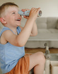 A smiling boy explores with a mushie Kaleidoscope while sitting on a cushioned chair.