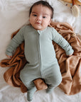 A baby wearing mushie Zipper Pajama in light green lies on a brown blanket atop a white bed, looking up with a slight smile and arms outstretched.