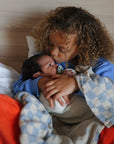 A young child with curly hair sits on a bed, lovingly holding and kissing a newborn wrapped in a mushie Organic Cotton Muslin Swaddle Blanket.