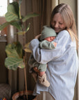 A woman in a striped shirt gently holds a sleeping baby wearing mushie Chunky Knit Pants, standing by a window with potted plants in a cozy, softly lit room.