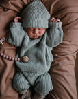 A newborn sleeps with arms raised on a brown cushion, wearing mushie Chunky Knit Pants in organic cotton and a green hat, next to a clipped pacifier with beaded string.