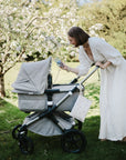 A woman in a long white dress smiles as she leans over a gray baby stroller in a garden, her mushie Water Resistant Wet Bag for diaper storage nearby. Blossoming trees frame the peaceful scene in the background.