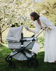 A woman in a long white dress smiles as she leans over a gray baby stroller in a garden, her mushie Water Resistant Wet Bag for diaper storage nearby. Blossoming trees frame the peaceful scene in the background.