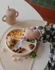 A toddler in a checkered long-sleeve bib uses a fork to eat pasta, corn, broccoli, and meat from a mushie Silicone Suction Plate. A beige sippy cup sits nearby, making baby feeding time easy and mess-free.