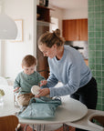 A woman in a light blue sweater packs a mushie Water Resistant Wet Bag on a kitchen table with green tiled walls, while her young child sits nearby reaching for something she holds. Natural light fills the room.