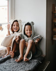 Two young children in hooded towels with bear ears sit by a window, smiling. One holds a fan, while the other explores the mushie Phone Press Toy to encourage fine motor skills. Stacks of books and toys are visible on the right.
