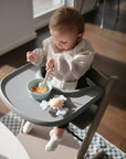 A baby sits in a high chair wearing the mushie Long Sleeve Bib, which is water resistant, while eating from a blue bowl. Sunlight streams through the windows, and a toy rests on the tray.