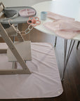 A Mushie Splat Mat lies under a modern high chair by a white table set with a pink cup and plate. A flower pacifier clip and pink silicone bib add color to the setup on the dark wooden floor.