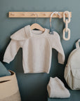 A beige knitted baby sweater hangs on a wooden peg rail, beside a mushie Silicone Pacifier Clip | Eva, large wooden chain links, folded knit pants, a beige backpack, and a woven basket filled with cushions.