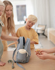 A woman with three children is by a table; the youngest, holding a FRIGG Daisy Silicone Pacifier, stands on the table as she opens a blue bag containing a water bottle. Soft light and toys create a cozy atmosphere.