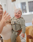 A smiling woman holds a laughing baby in green, happily chewing on a FRIGG Lucky Symmetrical Silicone Pacifier (0-6 Months), while a young boy in yellow stands nearby in a bright, cozy room with large windows.