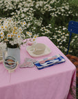 A table with a pink cloth, daisies, dishes, and a Mushie Bath Rinse Cup in a garden setting.