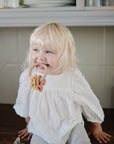 A smiling blond child in a white blouse sits on a kitchen counter, holding a mushie Nature Teething Ring in their mouth. Dishes and jars are displayed on the shelves behind them.