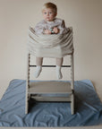 A toddler sits in a high chair with a light beige cover, holding a small object. The chair is on the Mushie Splat Mat, a blue water-resistant mat, against a plain tan background.