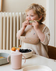 A young child with curly blond hair sits at a table, wearing a bib and eating melon. In front of them is the Mushie Trainer Sippy Cup with easy-grip handles, along with a bowl of grapes and other snacks.